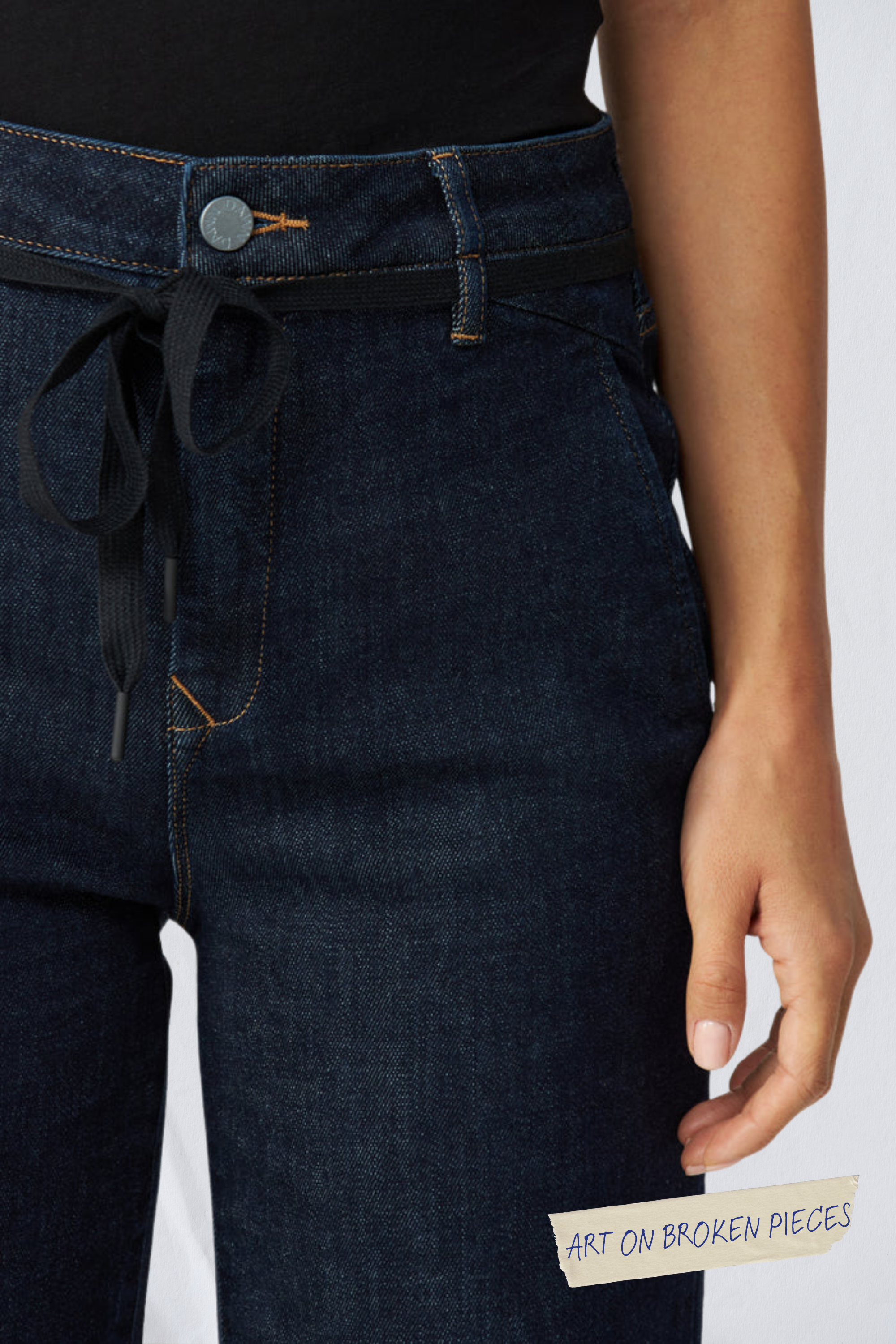 Dunkelblaue DEW Flared Denim mit French Pocket, hohem Bund, schwarzem Kordelzug und orangenen Nähten; am Bein gut sichtbar das Etikett 'ART ON BROKEN PIECES'.