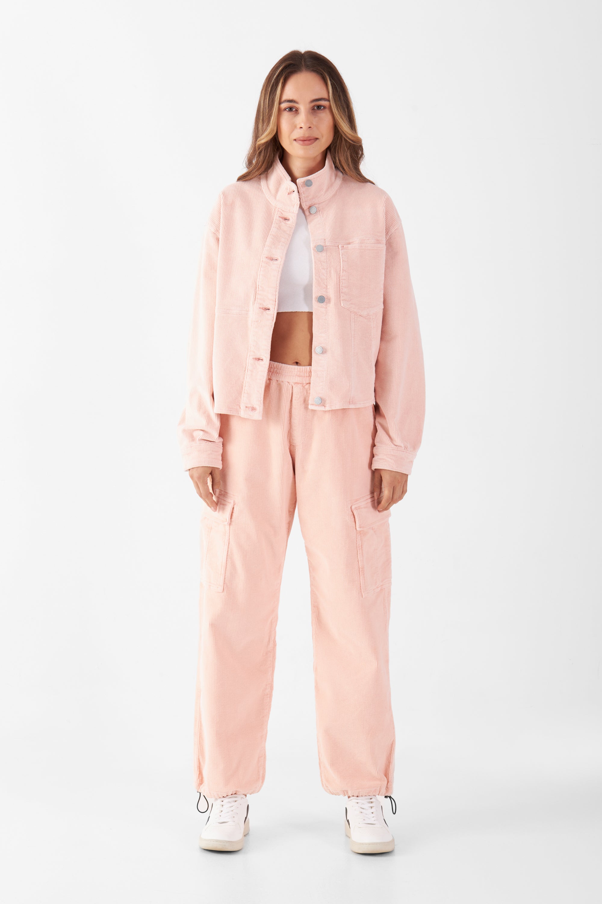 Rosa Cordhose mit Mid-Waist, elastischem Bund und Cargotaschen; passende Cordjacke im gleichen Ton; Modell trägt weiße Sneaker.